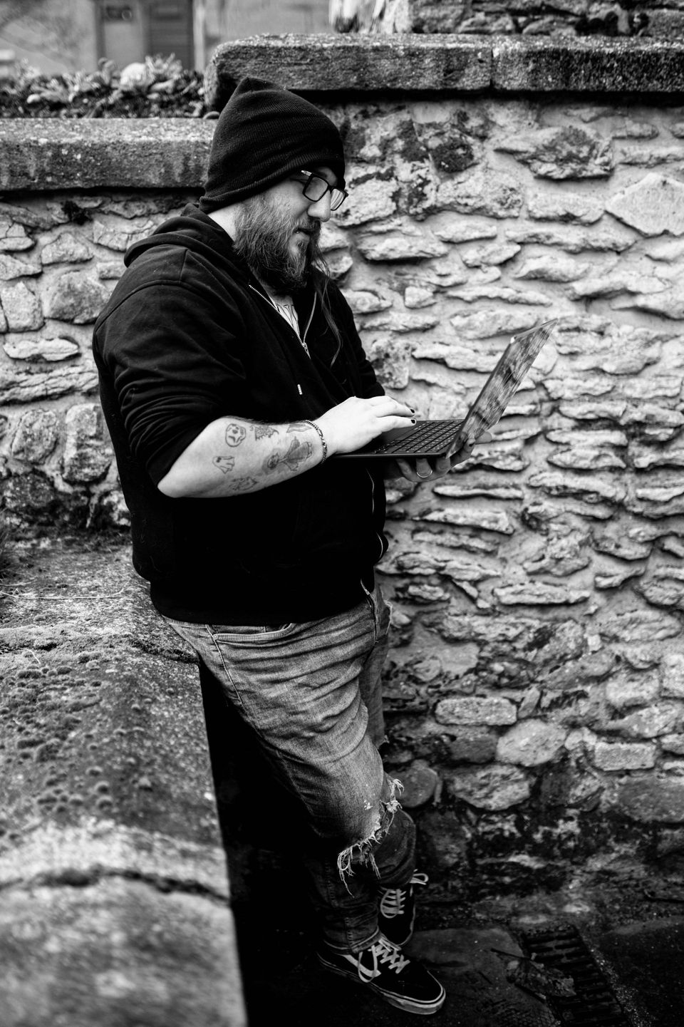 Nathan Pearson in a beanie and hoodie, leaning against a stone wall, typing on a laptop. Black and white.